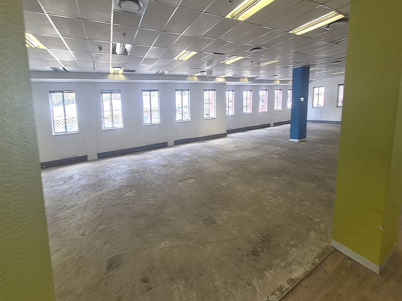 A large, open-plan office space featuring a suspended ceiling, fluorescent lighting, and multiple windows, currently awaiting flooring installation.