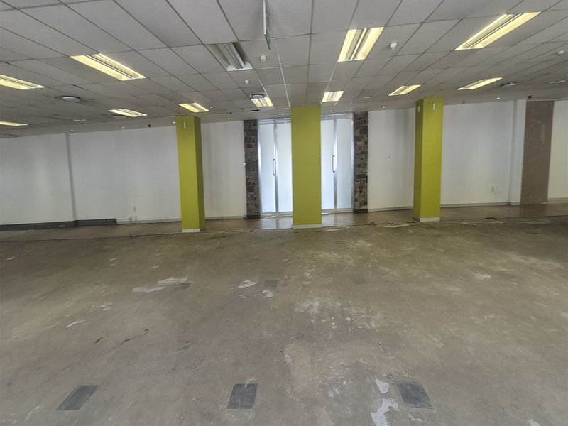 A large, vacant open-plan office space featuring a suspended ceiling, recessed lighting, and structural columns, currently requiring flooring refurbishment.