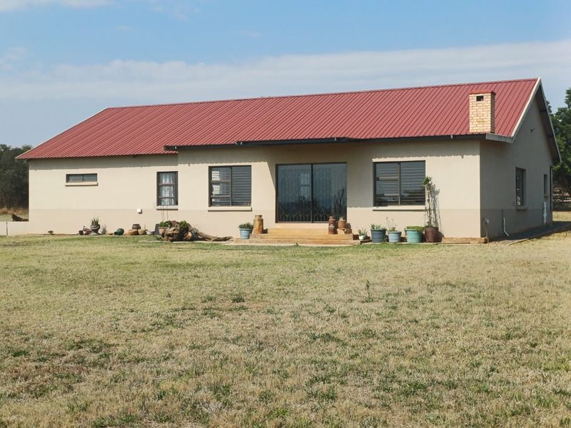 Rear view of a single-story farm house featuring a red corrugated metal roof, brick chimney, and sliding glass door access to the garden.