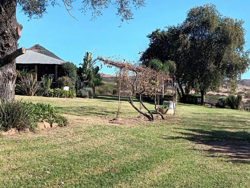 A spacious farm garden featuring mature trees, a well-maintained lawn, and a rustic trellis, with a view of the main dwelling and surrounding landscape.
