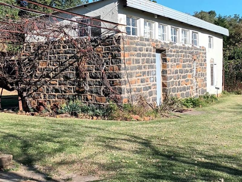 A rustic farm building featuring traditional stone masonry walls, a corrugated metal roof, and an attached metal trellis structure set within a grassy garden.