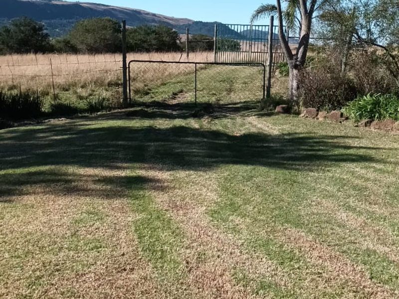 A view of a rural farm property featuring a metal gate, wire fencing, and expansive grass fields under a clear blue sky.