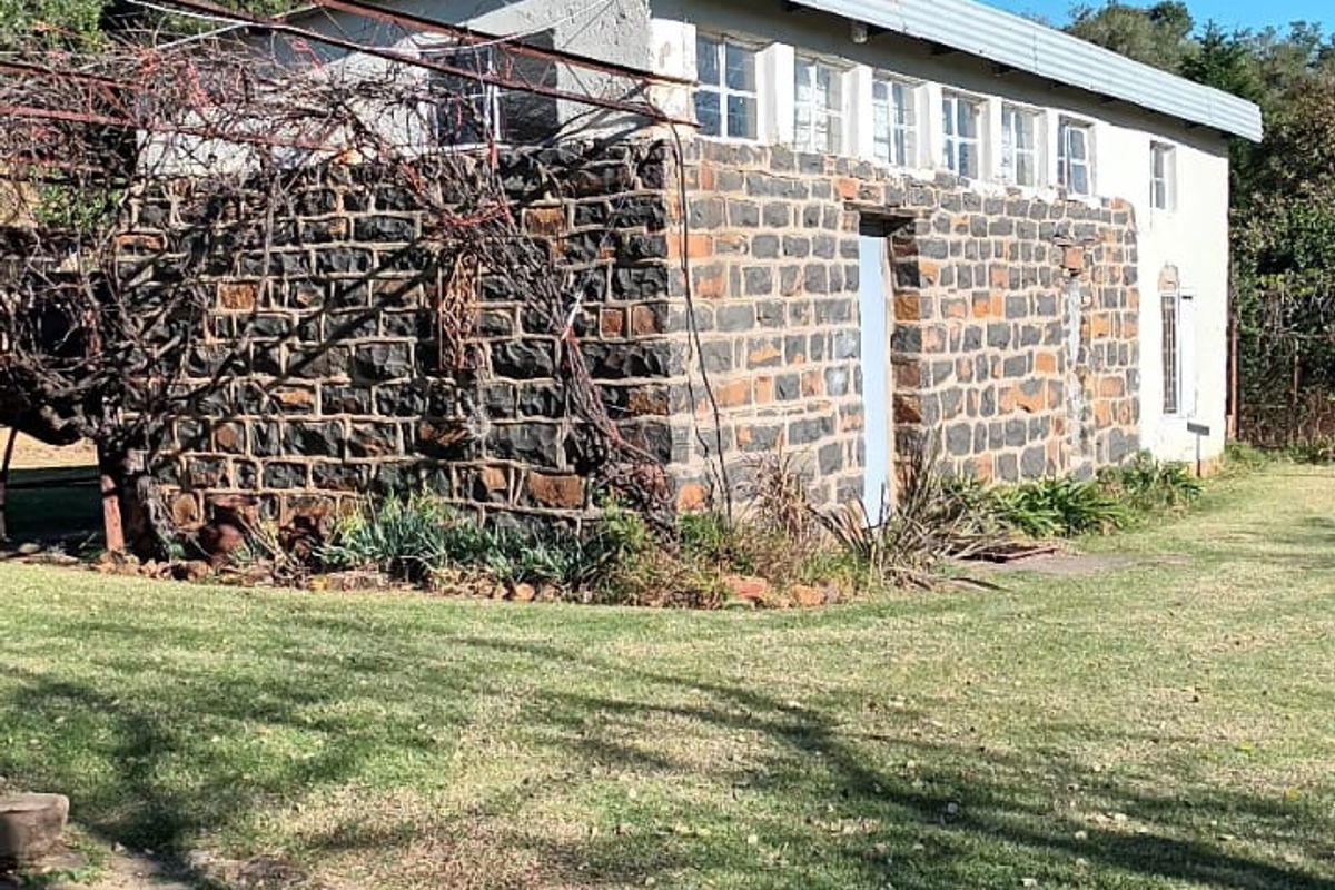 A rustic farm building featuring traditional stone masonry walls, a corrugated metal roof, and an attached metal trellis structure set within a grassy garden.