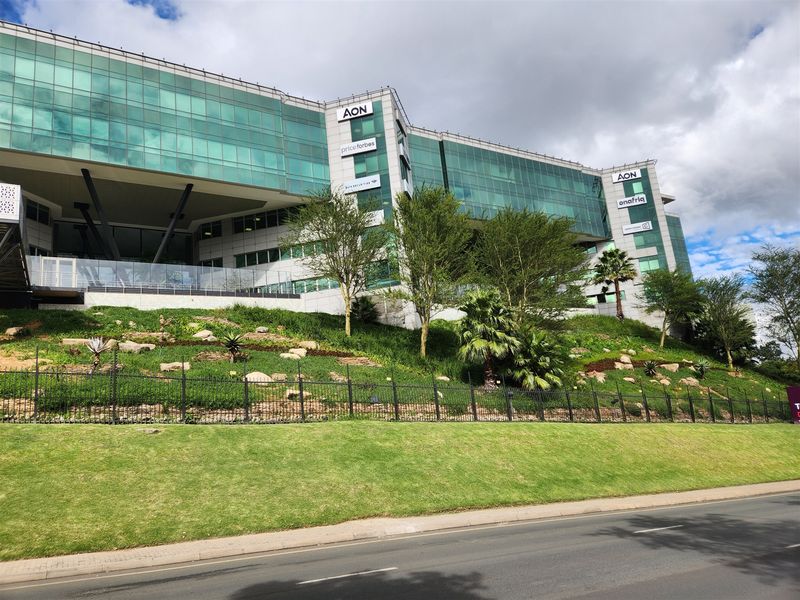 A modern multi-story office building featuring a glass facade, metal cladding, and a well-maintained landscaped embankment with perimeter fencing.