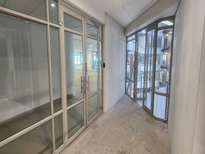 A bright, empty office hallway featuring glass partitions and doors, leading to an open-plan workspace with unfinished flooring and neutral wall finishes.