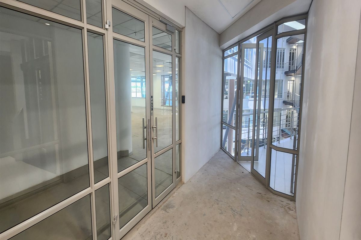 A bright, empty office hallway featuring glass partitions and doors, leading to an open-plan workspace with unfinished flooring and neutral wall finishes.
