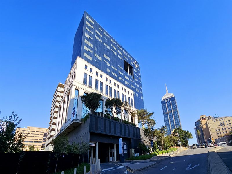 A modern high-rise office building featuring a glass curtain wall facade, integrated parking entrance, and elevated terrace landscaping in a prominent urban location.