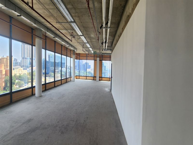 An expansive, unfinished office space featuring exposed industrial ceilings, structural columns, and floor-to-ceiling windows offering extensive city views.