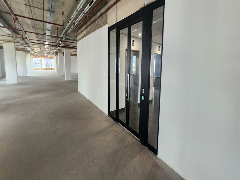 An open-plan office space in shell condition featuring exposed ceiling services, concrete flooring, and modern black-framed glass partitioning.