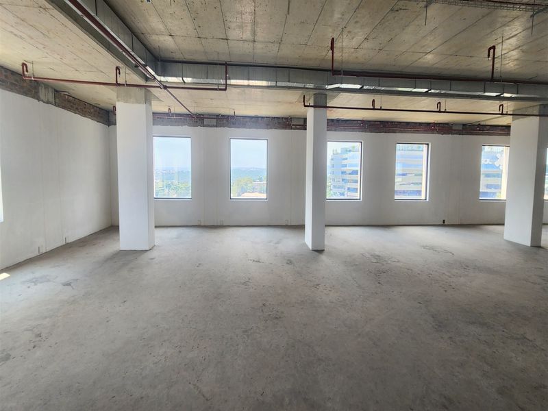 An open-plan office space in shell condition featuring exposed concrete ceilings, fire sprinkler systems, and large windows with natural light.