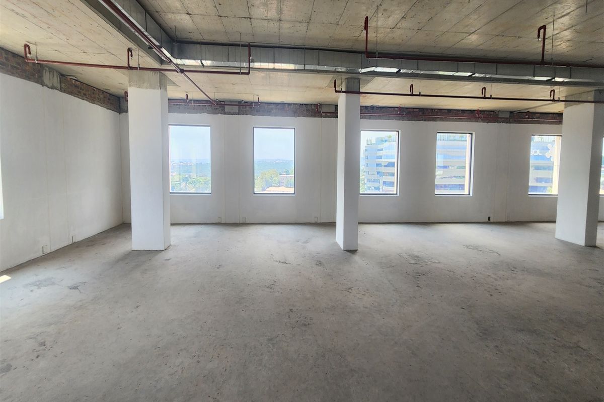 An open-plan office space in shell condition featuring exposed concrete ceilings, fire sprinkler systems, and large windows with natural light.