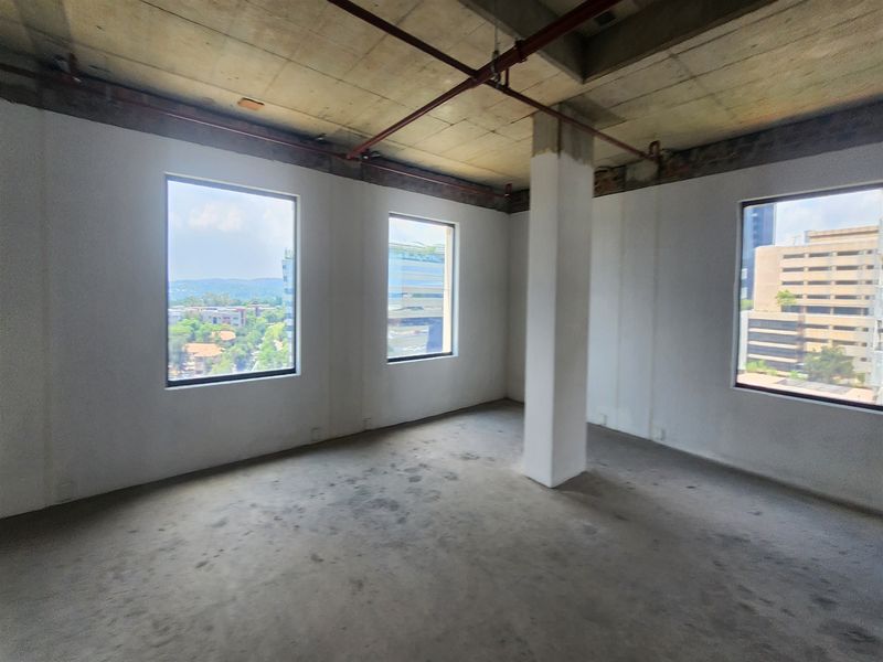 An unfinished open-plan office space featuring exposed concrete ceilings, industrial fire sprinkler piping, and large windows providing ample natural light and city views.