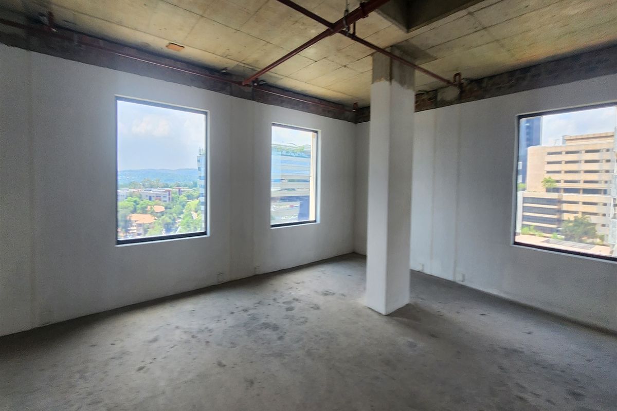 An unfinished open-plan office space featuring exposed concrete ceilings, industrial fire sprinkler piping, and large windows providing ample natural light and city views.
