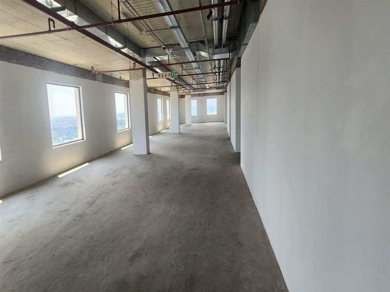An expansive, open-plan office space in shell condition, featuring exposed industrial ceilings, concrete floors, and large windows providing ample natural light.