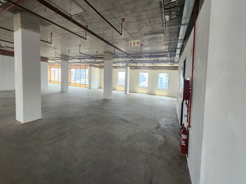 An expansive, open-plan office space featuring an industrial aesthetic with exposed concrete ceilings, structural columns, and integrated fire safety infrastructure.