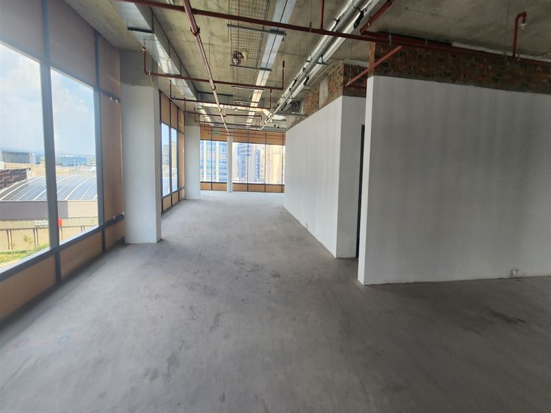 An open-plan office space in shell condition, featuring exposed industrial ceilings, concrete floors, and large windows with city views.