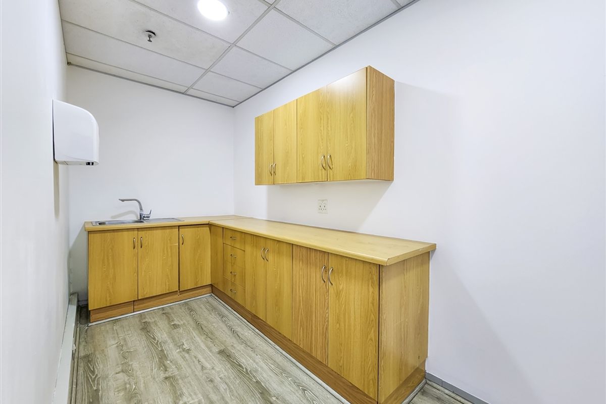 A clean, functional office kitchenette featuring wood-look laminate cabinetry, a stainless steel sink, and durable vinyl flooring under a drop ceiling.
