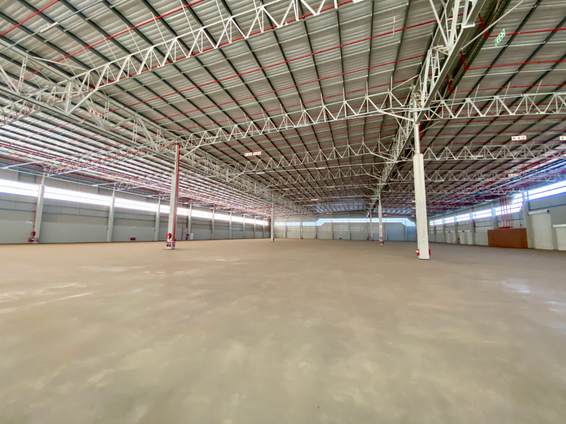 A vast, open-plan industrial warehouse space featuring high ceilings with exposed steel trusses, fire sprinkler systems, and polished concrete flooring.