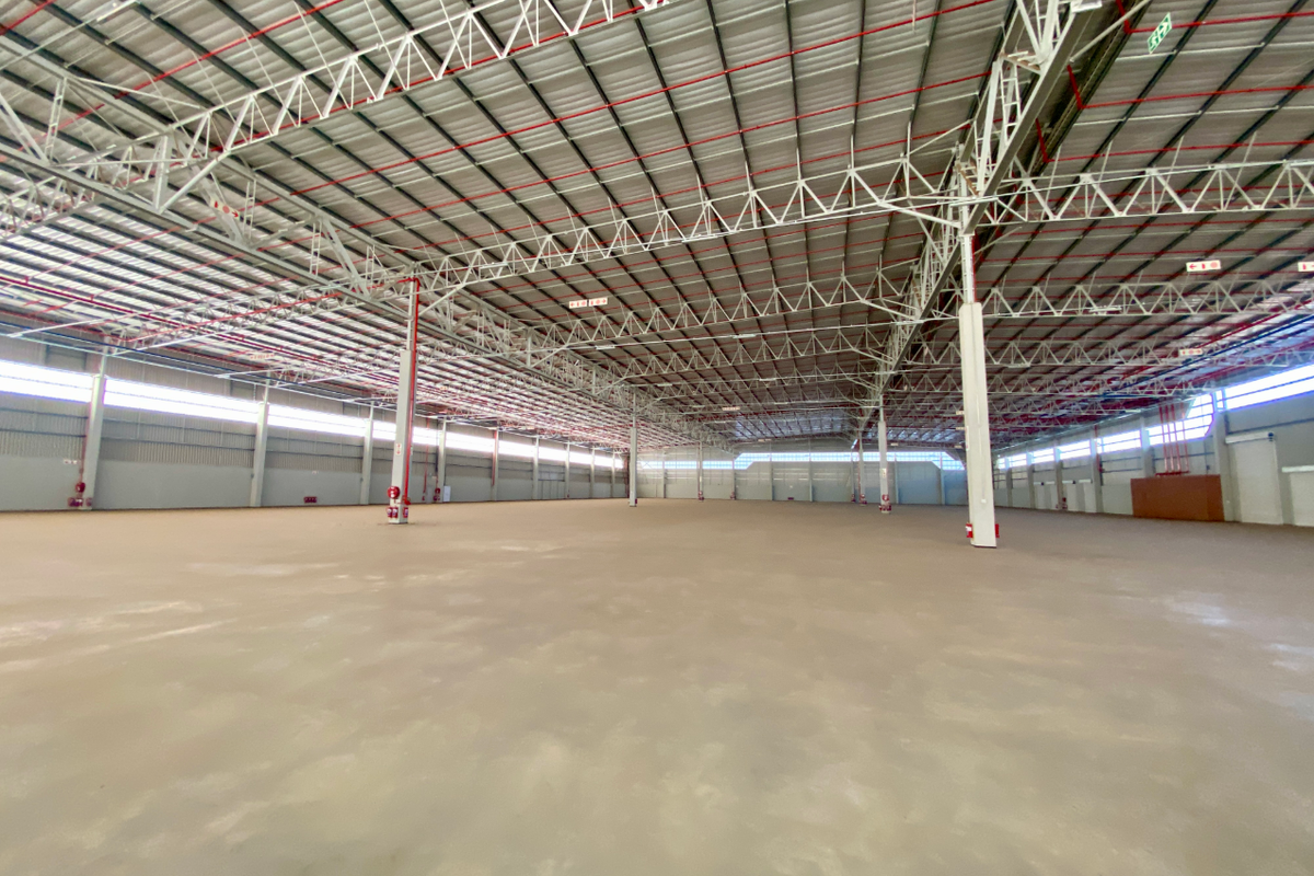 A vast, open-plan industrial warehouse space featuring high ceilings with exposed steel trusses, fire sprinkler systems, and polished concrete flooring.