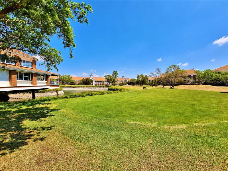 A scenic view of a well-maintained golf course green, water feature, and surrounding office park architecture under a clear blue sky.