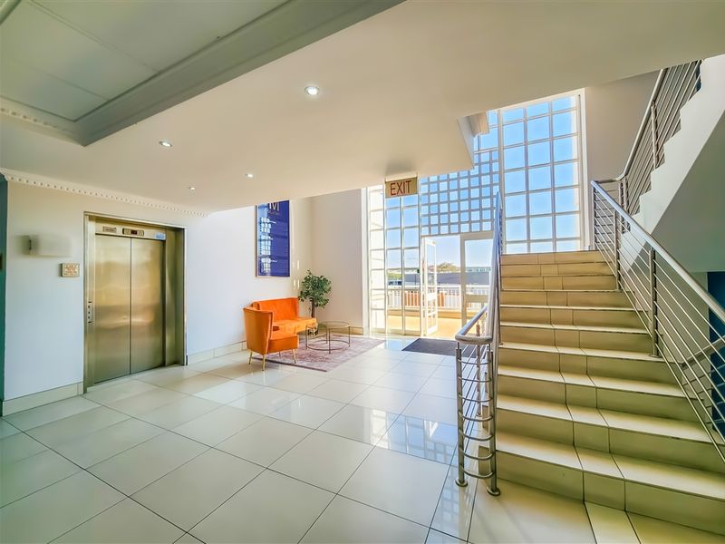 Bright and modern office building lobby featuring polished tile floors, a stainless steel elevator, and a prominent staircase with glass window accents.