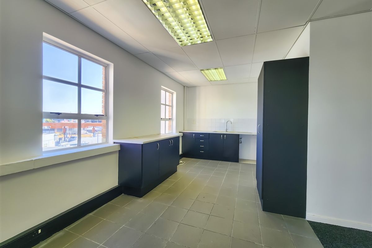 A functional office kitchenette featuring dark cabinetry, a sink, tiled flooring, and bright overhead lighting in a clean, empty space.