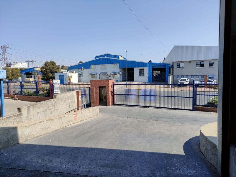 Front view of an industrial property featuring a secure gated entrance, concrete driveway, and warehouse facility with metal cladding.