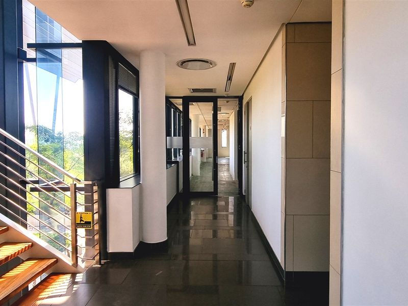 A modern, well-lit office hallway featuring tiled flooring, a sleek staircase with stainless steel balustrades, and large windows providing natural light.