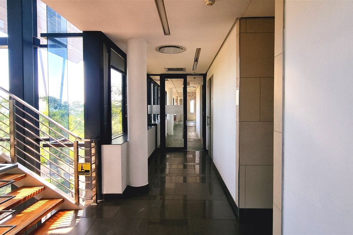 A modern, well-lit office hallway featuring tiled flooring, a sleek staircase with stainless steel balustrades, and large windows providing natural light.