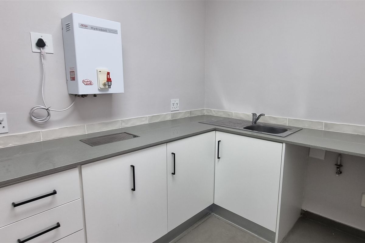 A clean, modern kitchenette featuring white cabinetry, stone countertops, a stainless steel sink, and a wall-mounted boiling water dispenser.