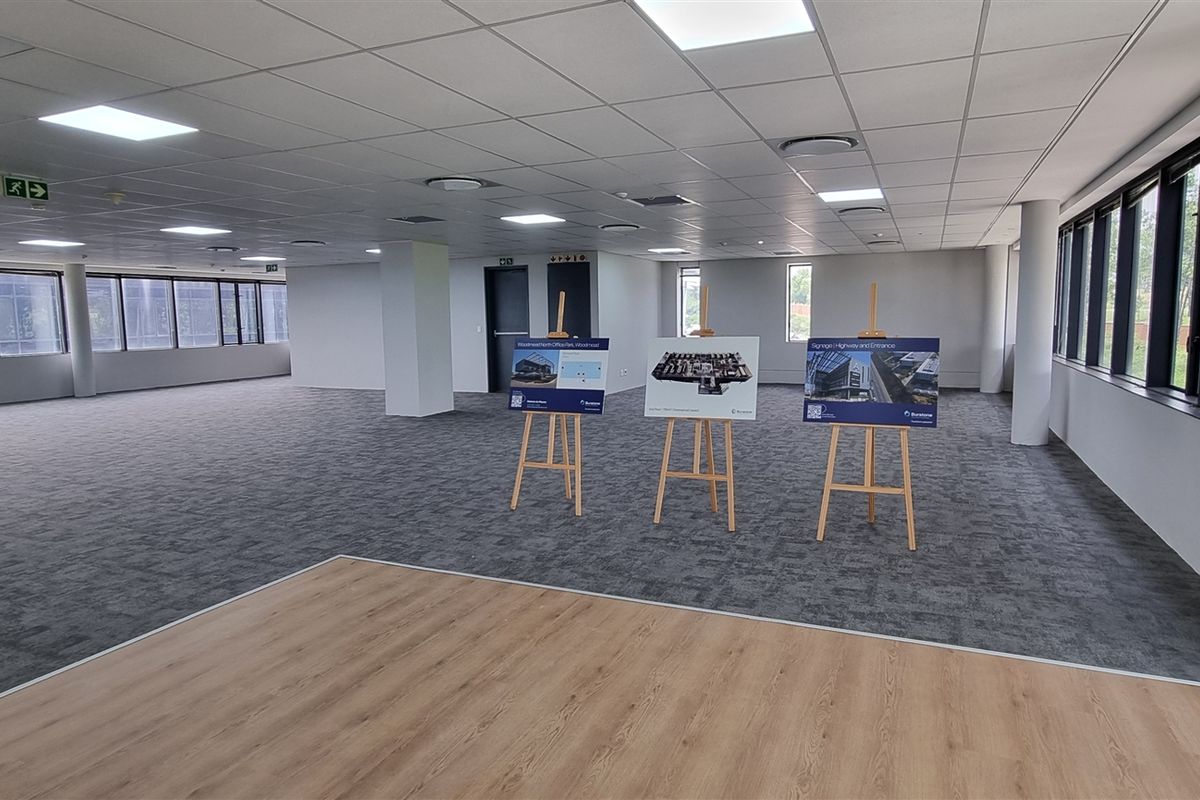 A spacious, open-plan office area featuring modern carpet tiles, laminate flooring, recessed lighting, and extensive windows providing ample natural light.