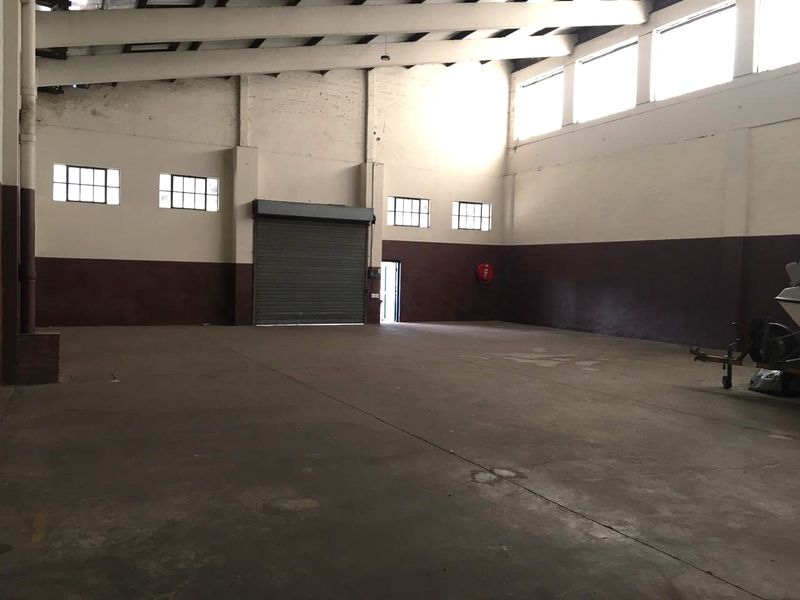 Spacious industrial warehouse unit featuring high ceilings with exposed steel trusses, concrete flooring, and a large roller shutter door for easy access.
