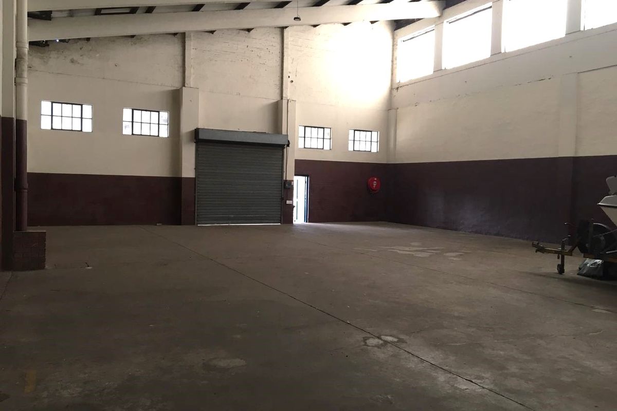 Spacious industrial warehouse unit featuring high ceilings with exposed steel trusses, concrete flooring, and a large roller shutter door for easy access.
