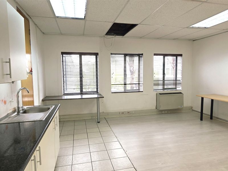 An empty office space featuring a small kitchenette with a sink, drop ceiling, HVAC unit, and large windows with venetian blinds.