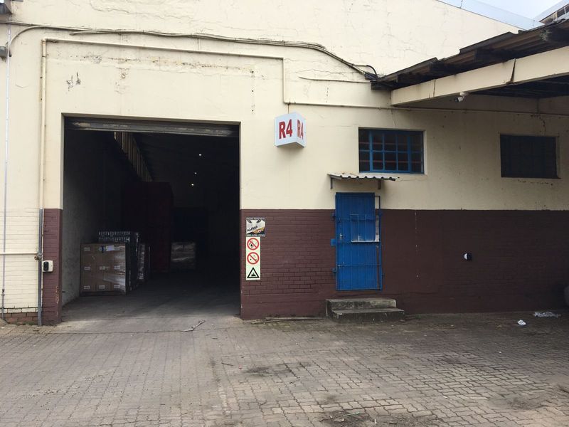 Industrial warehouse exterior featuring a large roller shutter access point, security gate, and paved loading area with visible masonry wear.