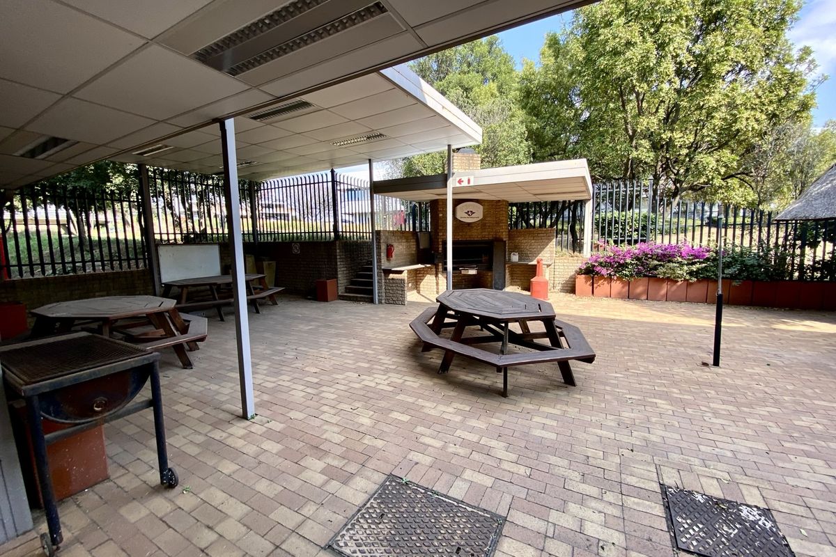 A spacious covered outdoor patio area featuring brick paving, a built-in braai, and perimeter security fencing, suitable for office staff recreation.
