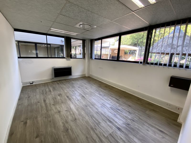 A bright, empty office space featuring wood-look vinyl flooring, a suspended ceiling with recessed lighting, and large windows providing natural light and garden views.