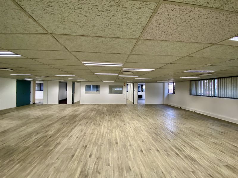 Spacious open-plan office area featuring modern laminate wood flooring, a suspended acoustic ceiling with recessed lighting, and large windows with vertical blinds.