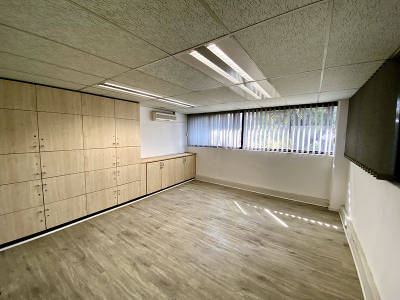 A bright, empty office space featuring extensive built-in storage cabinetry, wood-look laminate flooring, recessed lighting, and a wall-mounted air conditioning unit.