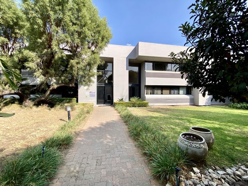 A well-maintained office building exterior featuring a paved walkway leading to a modern glass entrance, surrounded by established landscaping and mature trees.