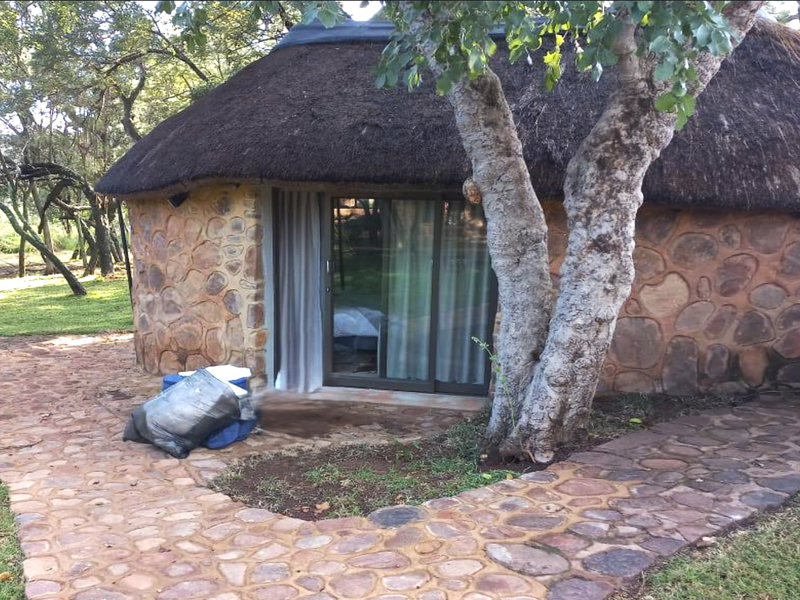 A rustic round cottage featuring traditional thatch roofing, stone exterior cladding, and a sliding glass door entrance set within a natural garden landscape.