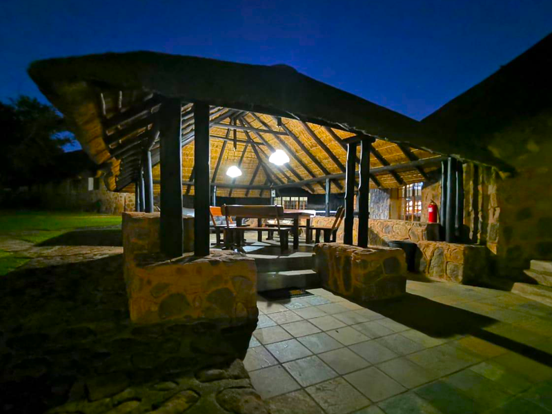 A covered outdoor patio area featuring a traditional thatched roof, stone masonry pillars, and durable tiled flooring, perfect for evening outdoor entertainment.