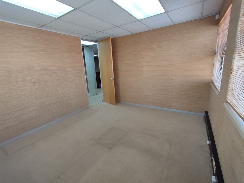 A vacant office space featuring wood veneer wall paneling, suspended ceiling with recessed lighting, and carpet flooring with visible wear.