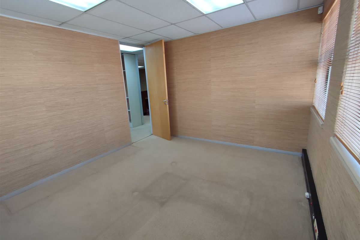 A vacant office space featuring wood veneer wall paneling, suspended ceiling with recessed lighting, and carpet flooring with visible wear.