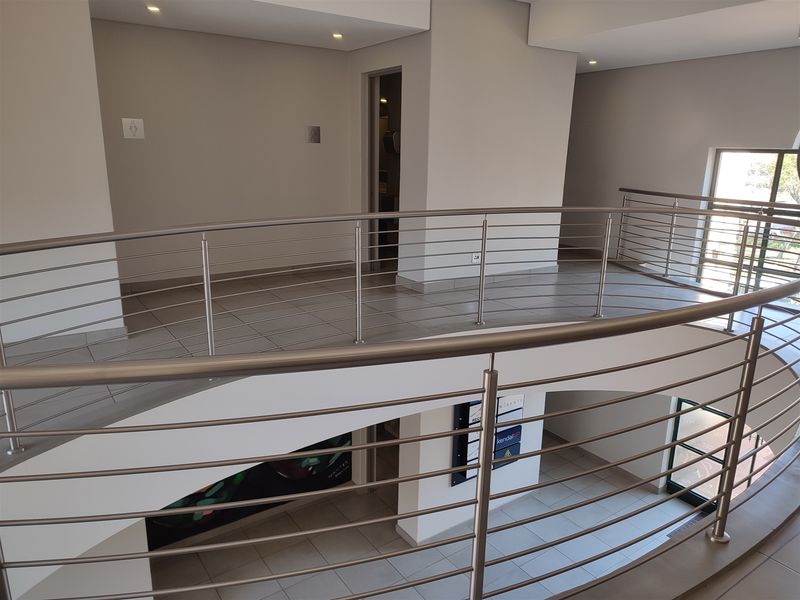 A modern office hallway featuring tiled flooring, a sleek stainless steel balustrade overlooking the lower level, and clean, neutral wall finishes.