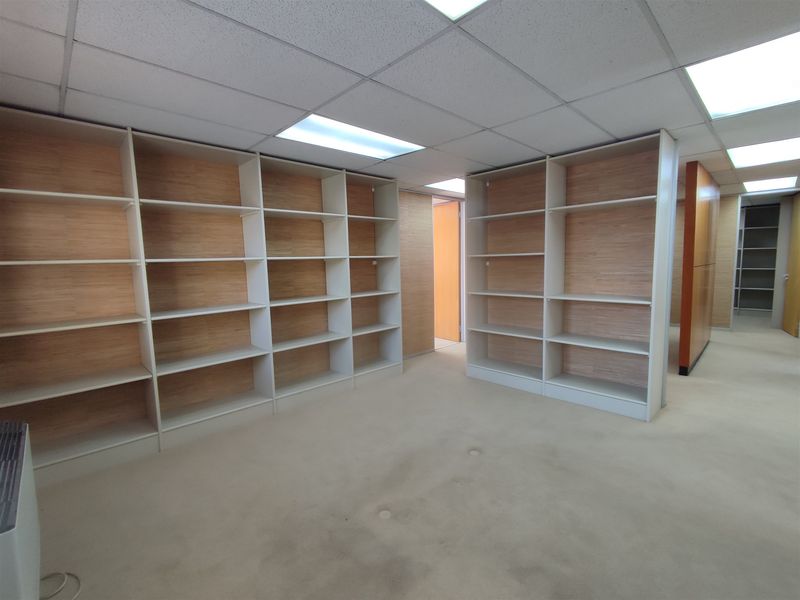 An empty office space featuring extensive built-in wooden shelving units, a suspended ceiling with recessed lighting, and neutral carpet flooring.