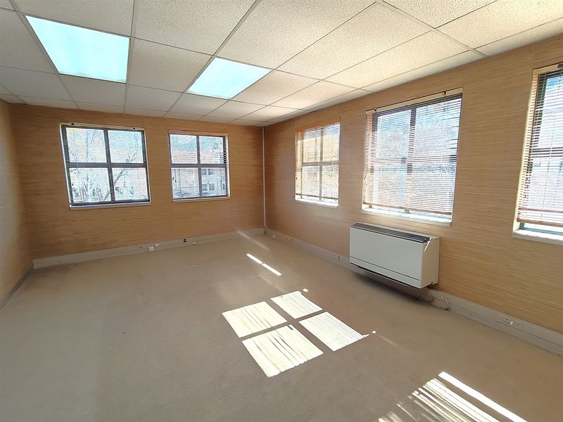 A bright, empty office space featuring suspended ceilings, recessed lighting, carpeted flooring, and a wall-mounted HVAC unit with large windows.