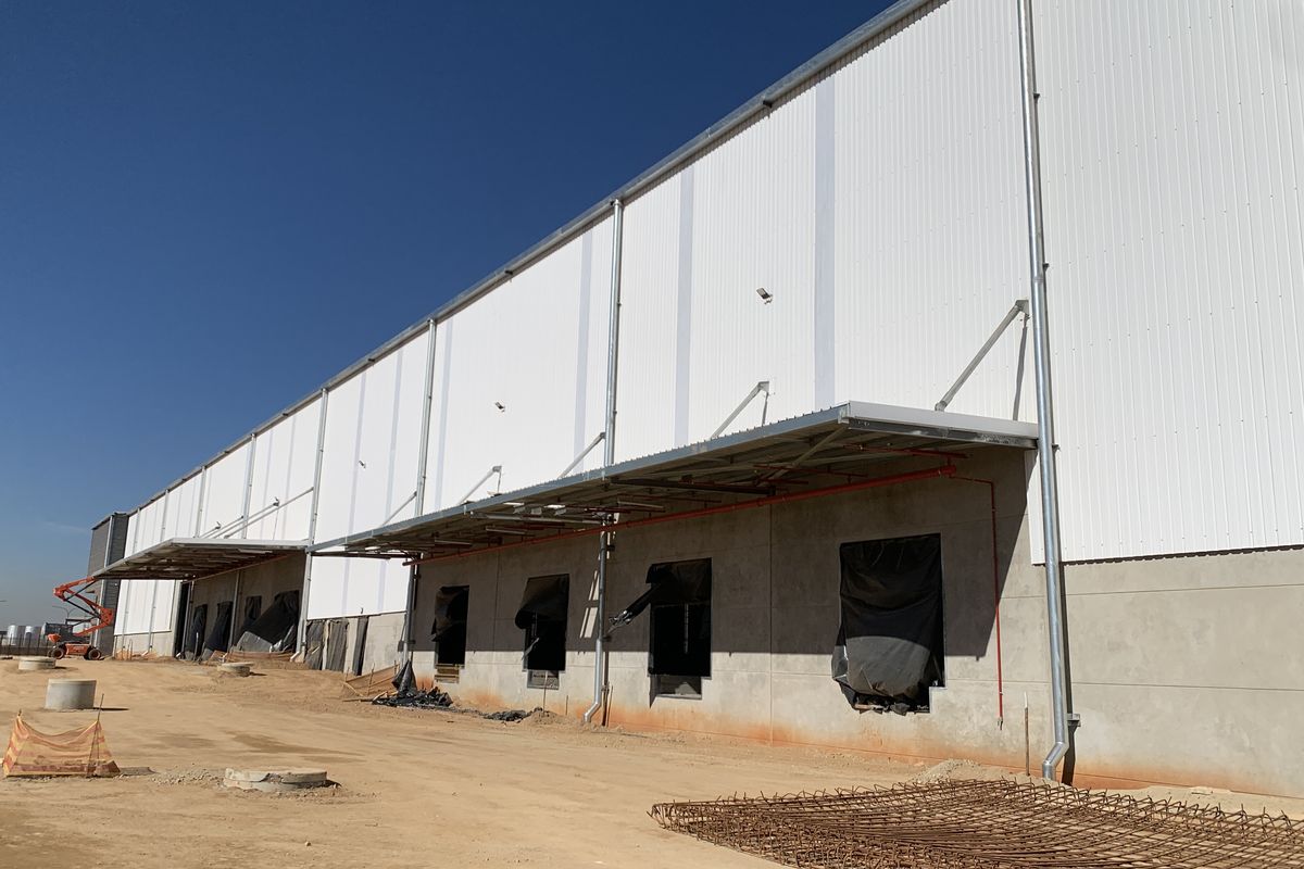 Exterior view of an industrial warehouse facility featuring corrugated metal cladding, loading bay canopies, and concrete base walls under construction.