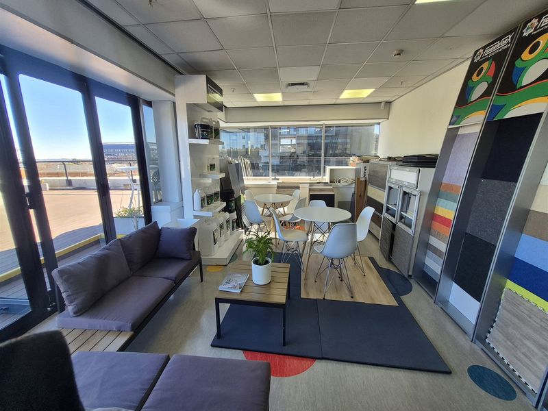 A bright office space featuring large windows, sliding glass doors leading to an outdoor area, suspended ceilings, and integrated product display shelving.