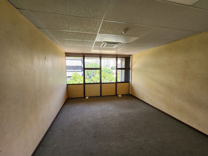 An empty office space featuring a suspended ceiling with integrated air conditioning, large windows providing natural light, and carpeted flooring.